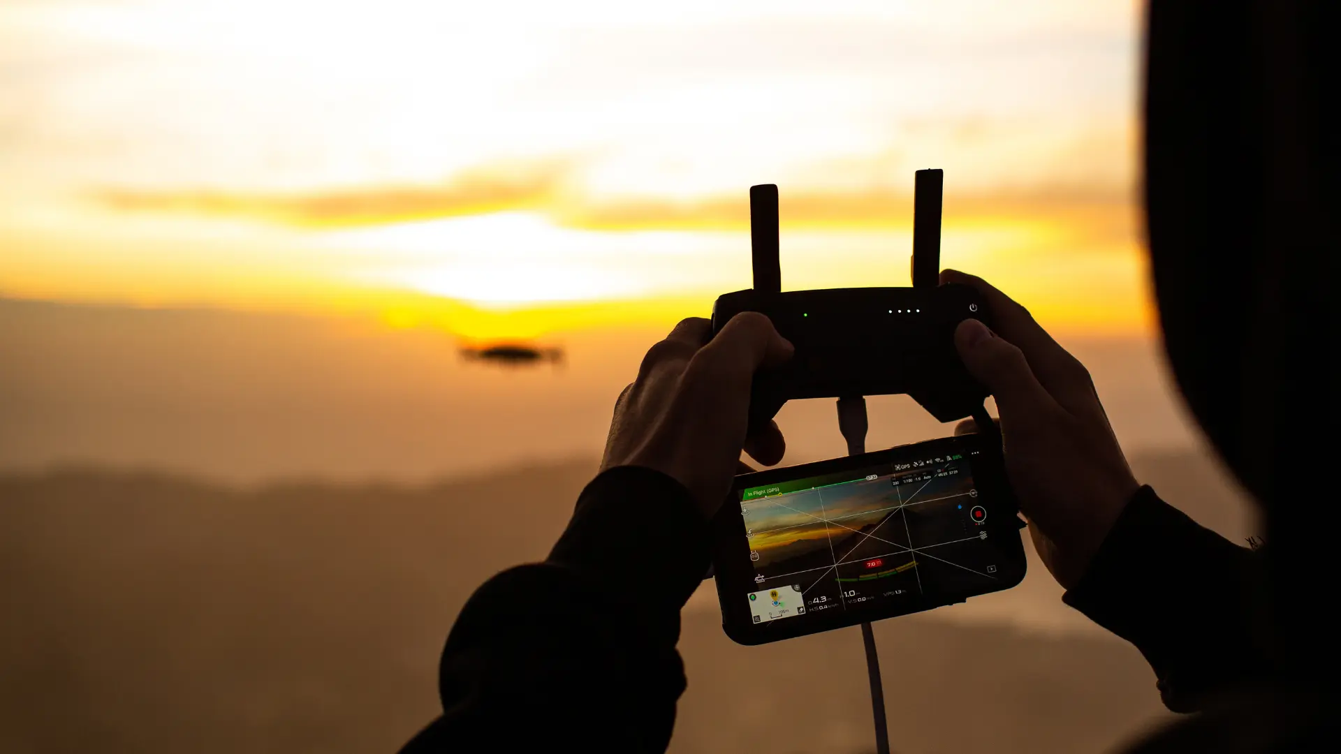 Close-up of a remote pilot holding a controller and smartphone to monitor an aerial videography session at sunset.